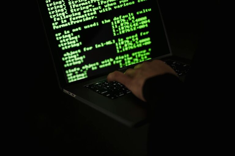 Crop unrecognizable computer geek typing on netbook with codes on screen while hacking system in darkness